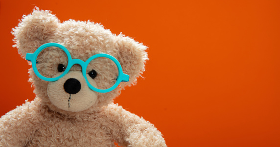 Back to school, eyesight test. Smart kid, cute teddy wearing blue eyeglasses against orange color background, copy space.