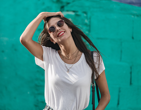 Cheerful young woman in white t shirt and golden necklace touching long hair smiling and looking at camera on turquoise wall background