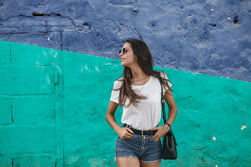 Carefree stylish woman in casual wear standing by colorful blue and turquoise grunge wall and looking away on daytime