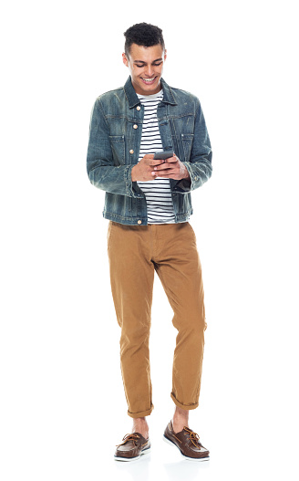 Looking down / full length / front view of 20-29 years old adult handsome people / tall person african ethnicity / african-american ethnicity male / young men standing in front of white background wearing boat shoe / denim jacket who is smiling / happy / cheerful / cool attitude and holding mobile phone / using smart phone / text messaging