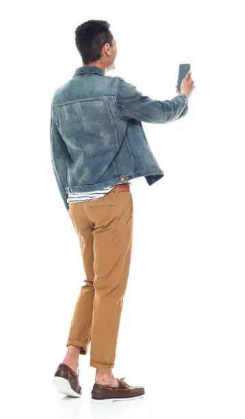 Full length / rear view of 20-29 years old adult handsome people / tall person african ethnicity / african-american ethnicity male / young men standing in front of white background wearing boat shoe / denim jacket / cool attitude who is taking a selfie / photographing and holding mobile phone / using smart phone