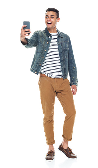 Full length / front view of 20-29 years old adult handsome people / tall person african ethnicity / african-american ethnicity male / young men standing in front of white background wearing boat shoe / denim jacket who is smiling / happy / cheerful / cool attitude who is taking a selfie / photographing and holding mobile phone / using smart phone
