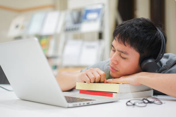 primo piano giovane asiatico che indossa cuffie wireless per studiare il corso online e dormire sopra il libro di testo dopo la fine, l'assistenza sanitaria e il concetto di persone lifestyle - sleeping high school desk education foto e immagini stock