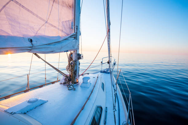 穏やかな水白いスループは日没時に航海するヨットを装備した。デッキから弓、マストおよび帆への眺め。バルト海, ラトビア - セーリング 写真 ストックフォトと画像