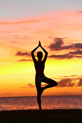 Yoga woman training in sunset in tree pose meditating outdoors by beach ocean sea. Female yoga instructor working out training in serene ocean landscape. Silhouette of woman model against sun.