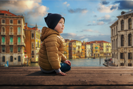Little boy sits on a wooden pier at sunset. Beautiful Venice on the background.