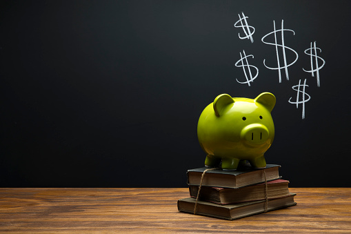 This is a photograph of a green Piggy Bank sitting on a desk a with a chalk drawing of Dollar Bill signs on Black Chalkboard Background. This image could relate to saving for a college fund and education.