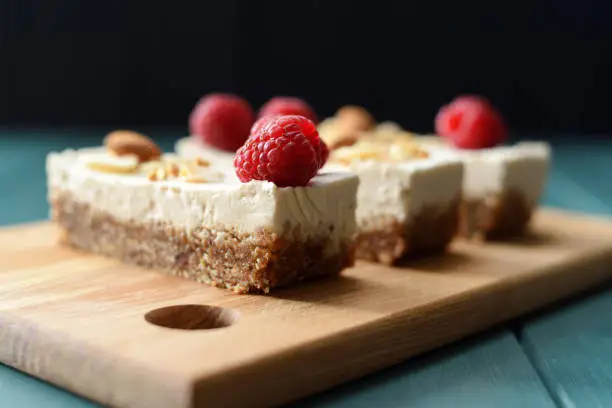 Vegan sugar free dessert. Coconut milk cheesecake bars decorated with raspberries, almonds and coconut flakes on dark background side view copy space