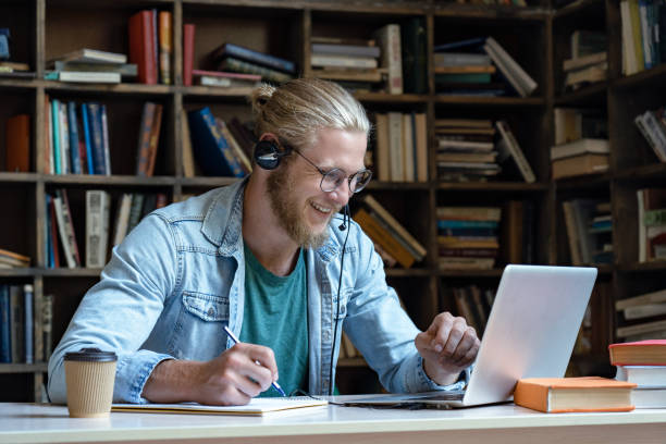 o olhar sem fio feliz de sorriso do homem novo do desgaste dos auriculares na tela do portátil faz anotações estudo e aprendizagem na biblioteca assistir webinar treinamento online curso de vídeo chamada, educação a distância, skype conceito de ensi - video conference camera - fotografias e filmes do acervo