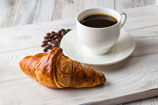 A white Cup of coffee with a croissant on a light wooden tray