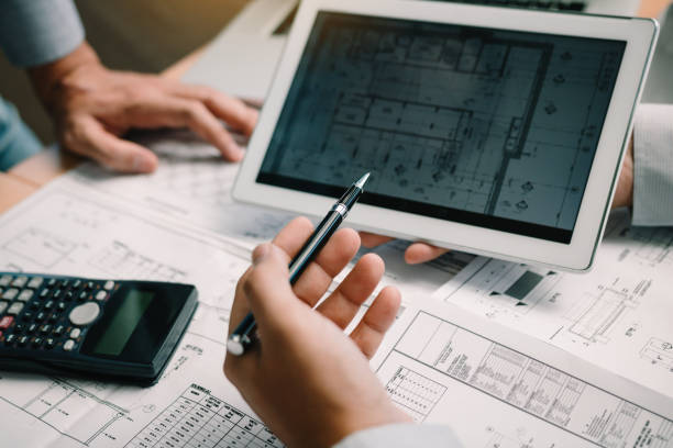 Two engineering working together and using digital tablet looking blueprint and analysis with architectural plan on desk. stock photo