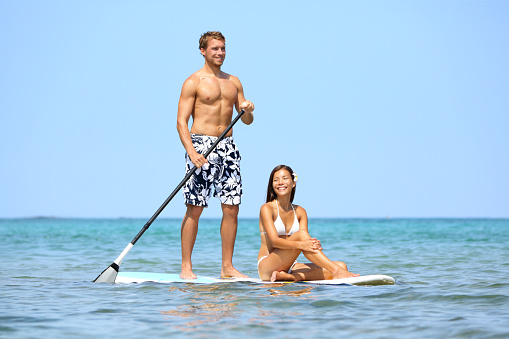 Beach fun couple on stand up paddleboard surfboard surfing together in ocean sea on Big Island, Hawaii Beautiful young multi-ethnic couple, mixed race Asian woman and Caucasian man doing water sport.