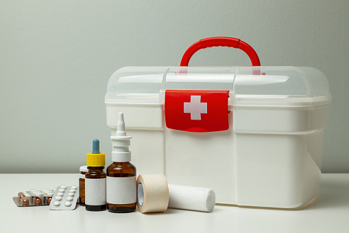 First Aid Kit. White box with a cross and a red fastener and pills with medicine bottles on a gray background.