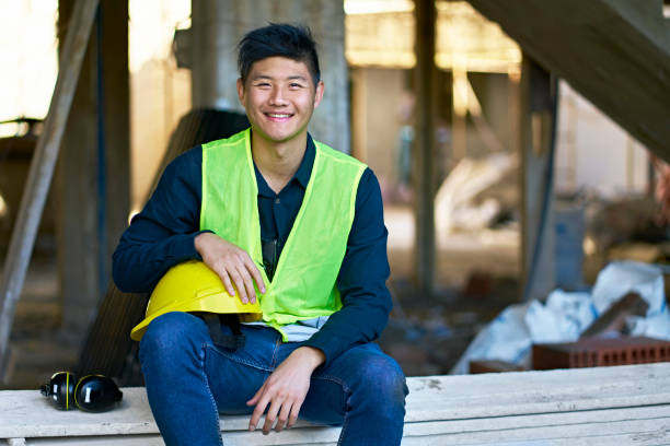 Portrait of smiling young building contractor sitting on planks. Male engineer is at construction site. He is wearing protective workwear.