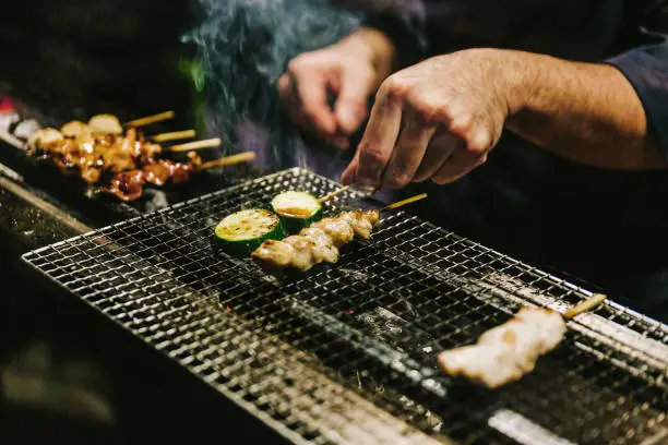 Close-up hands of Japanese Yakitori Chef grilling chicken marinated with ginger, garlic and soy sauce and cucumber with a lot of smoke.