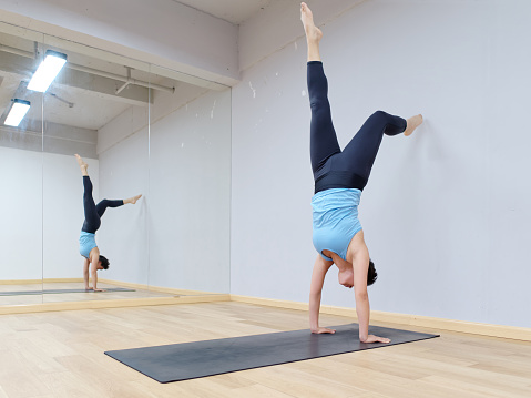 Young sporty attractive woman practicing yoga doing handstand pose working out wearing sportswear blue top and black pants on mat with mirror in yoga classroom, indoor full length portrait.