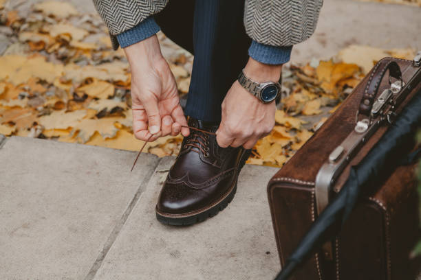 Gentleman is tying lace on brown shoe. Nearby is carpetbag Hands tie a thin lace on a stylish left brogue shoe in autumn park close-up. Nearby is a brown leather carpetbag brogue photos stock pictures, royalty-free photos & images