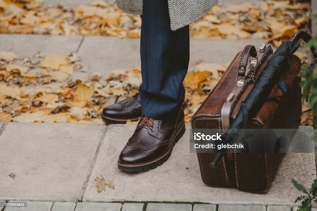 Legs of man and carpetbag in autumn park gentleman in the autumn square. Legs in brown brogue shoes stand on the step. Nearby is a leather antique carpetbag and umbrella cane. Brogue Stock Photo