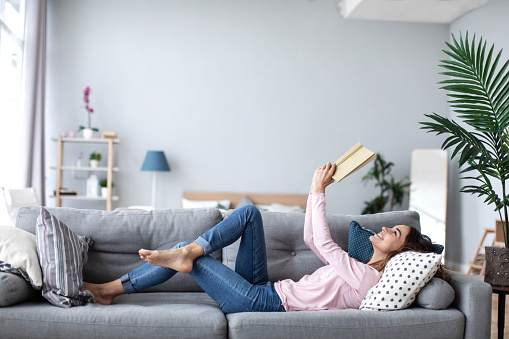 Beautiful smiling woman reading a book and lying on the sofa in the living room. Have a good time at home.