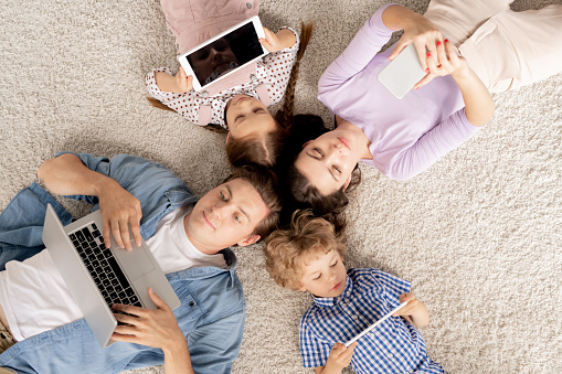 Top view of young family of four lying on the floor and scrolling in mobile gadgets while resting at home