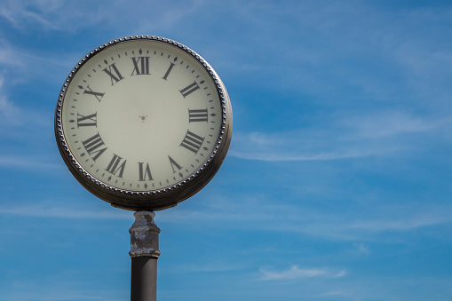 Old fashioned street clock with Roman numerals without arrows on blue slightly cloudy sky background. Real view of street clock in city. Stopped time concept.