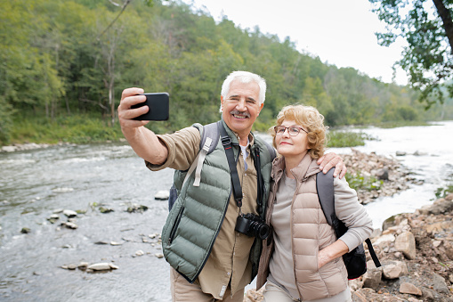 Happy mature active spouses looking at smartphone camera while making selfie by forest river during trip