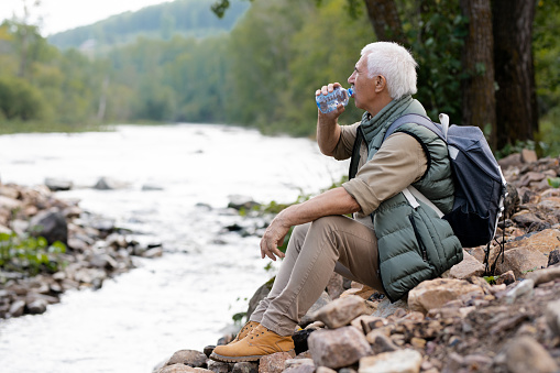 Tired mature backpacker drinking water from plastic bottle while sitting on river bank in natural environment