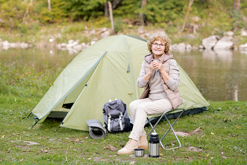 Mature smiling woman in activewear holding hot drink while sitting on small tourist chair by tent on river bank