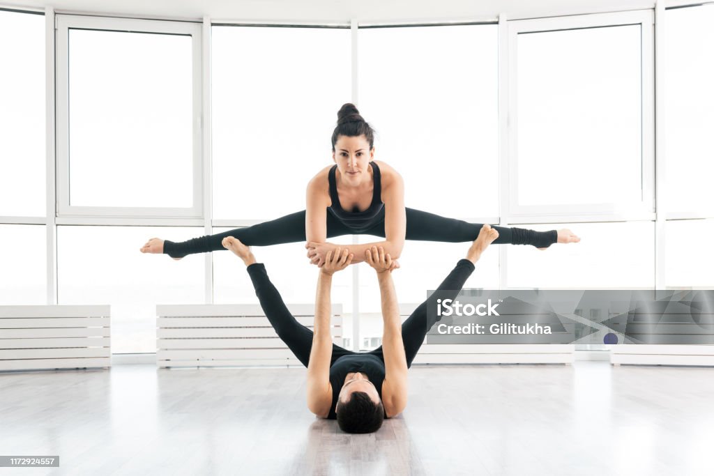 Young couple doing acro yoga in pair at studio Beautiful young man and woman practice pair acro yoga exercise in white fitness studio together with big windows on background. Man lying on wood floor and balancing womann in his feet Acroyoga Stock Photo