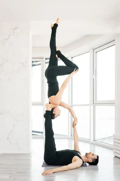 Beautiful young man and woman practice pair acro yoga exercise in white fitness studio together with big windows on background. Man lying on wood floor and balancing womann in his feet