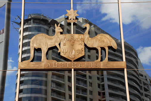 The Australian Coat of Arms in metal hangs on the glass wall of a building that reflects another high-rise across the street.