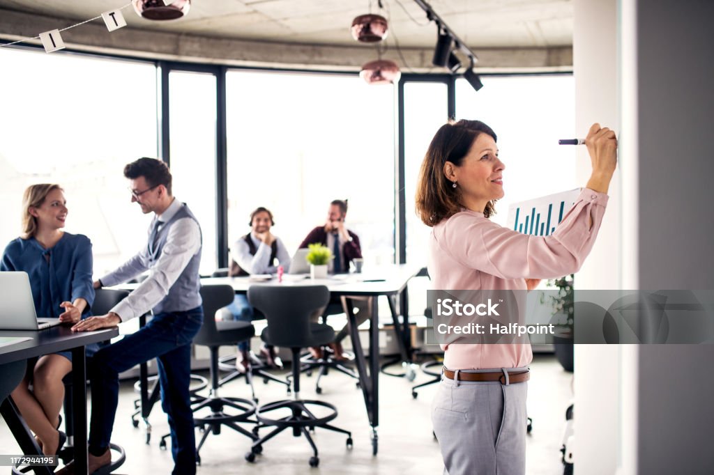 Businesspeople working in an office, writing on whiteboard. A portrait of businesswoman with colleagues standing in an office, writing on whiteboard. Adult Stock Photo