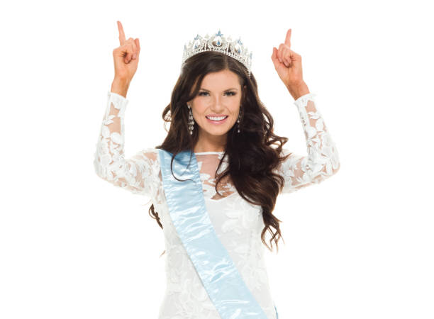 Waist up / looking up of 20-29 years old adult beautiful brown hair / long hair caucasian female / young women beauty queen / miss universe pageant in front of white background wearing crown / sash / dress who is success / winner / smiling / happy / cheerful who is pointing / beauty contest / glamour cintura hacia arriba / mirando hacia arriba de 20-29 años de edad adulto hermoso cabello castaño / pelo largo mujer caucásica / joven mujeres reina de belleza / miss universo desfile delante de fondo blanco usando corona / faja / vestido que está apunt