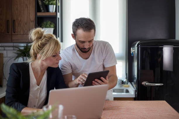 istock Couple in kitchen. Man is working on tablet while woman is working on laptop. 1172956070