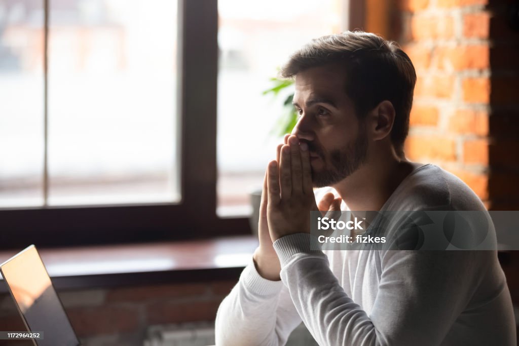 Concerned serious doubtful man sitting near laptop and thinking Young concerned businesswoman sitting at table near laptop folding hands together feels doubt unsure thinking making decision or waiting message good news having strong wish hope to get or win concept Decisions Stock Photo