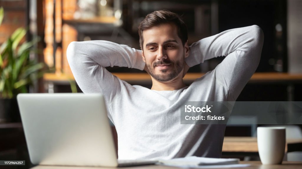 Serene businessman sitting at table feels satisfied accomplishing work Caucasian businessman sitting at table in cafe modern cozy office looking at laptop screen feels satisfied proud with done work, serene man resting putting hands behind head relaxing no stress concept Men Stock Photo
