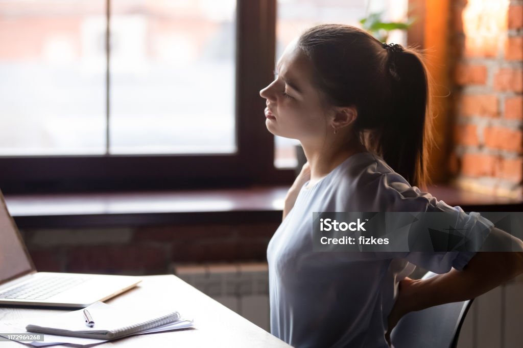 Female sitting at table after sedentary work feels back ache Side view young woman sitting at table working using computer take break touching massaging lower back feels discomfort after long sedentary studying, poor posture, incorrect stooped position concept Working Stock Photo