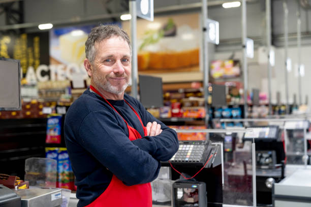 porträt von erwachsenen männlichen kassierer im supermarkt mit blick auf kamera lächelnd mit den armen gekreuzt - supermarket sales clerk retail cashier stock-fotos und bilder