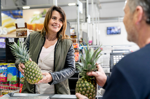 Female customer at checkout handing fresh pineapples to friendly sales clerk both smiling