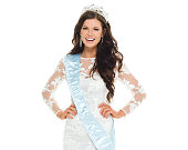 Waist up of 20-29 years old adult beautiful brown hair / long hair caucasian female / young women beauty queen / miss universe pageant in front of white background wearing crown / sash / dress who is success / winner / smiling / happy / cheerful