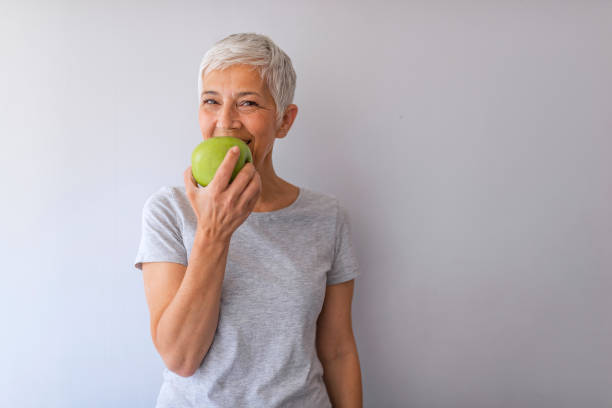 緑のリンゴを食べる灰色の壁の上の美しい先輩女性。 - women eating fruit food ストックフォトと画像