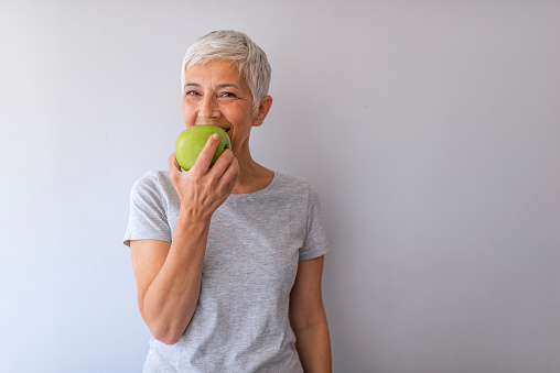 Portrait of happy mature woman holding granny smith apple at home. Beautiful senior woman over grunge grey wall eating green apple with happy face smiling.