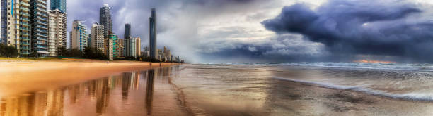 Stormy rainy clouds over Pacific ocean at sunrise moving towards city front of Surfers Paradise city CBD off sandy beach on Australian Gold Coast in wide panorama.