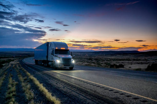 istock White Semi-Truck Speeding down a four lane highway with a dramatic sunset in the background and headlights on 1173038962