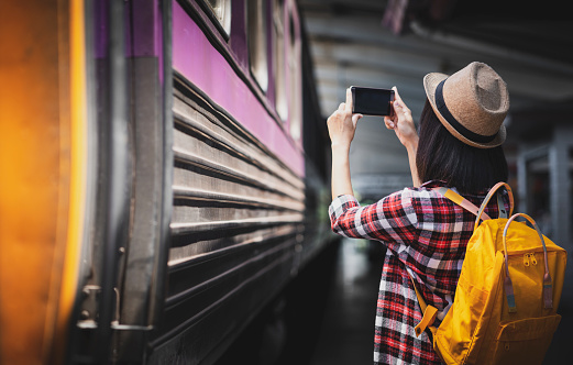 Tourist woman taking a photo with smartphone of side of train on railway background. Young asian woman traveler with backpack in the railway, Travel concept
