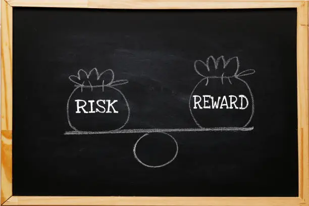 Photo of Write symbol Risk and reward bags on a basic balance scale in equal position on blackboard. risk management concept, depicts investors use a risk reward ratio to compare the expected return of an investment