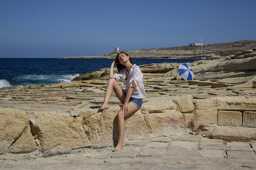 Young pretty woman in a white linen shirt and denim shorts on the windy rocky coast, enjoying summer vacation on the remote location far away from the tourist attractions