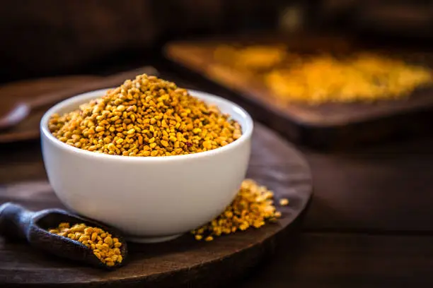 Front view of a bee pollen bowl on a rustic wooden table. Alongside the bowl is a wooden serving scoop filled with bee pollen and are at the left lower corner. At the right top corner is a defocussed wooden cutting boar with quinoa seeds on top. Low key DSLR photo taken with Canon EOS 6D Mark II and Canon EF 24-105 mm f/4L