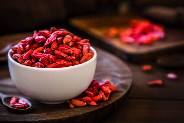Front view of a wolfberry bowl on a rustic wooden table. Alongside the bowl is a wooden serving scoop filled with wolfberry and are at the left lower corner. At the right top corner is a defocussed wooden cutting boar with quinoa seeds on top. Low key DSLR photo taken with Canon EOS 6D Mark II and Canon EF 24-105 mm f/4L