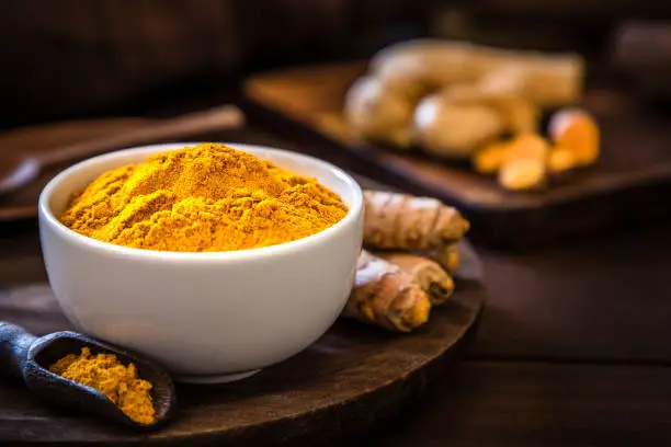 Front view of a turmeric powder bowl on a rustic wooden table. Alongside the bowl is a wooden serving scoop filled with turmeric powder and are at the left lower corner. At the right top corner is a defocussed wooden cutting boar with quinoa seeds on top. Low key DSLR photo taken with Canon EOS 6D Mark II and Canon EF 24-105 mm f/4L